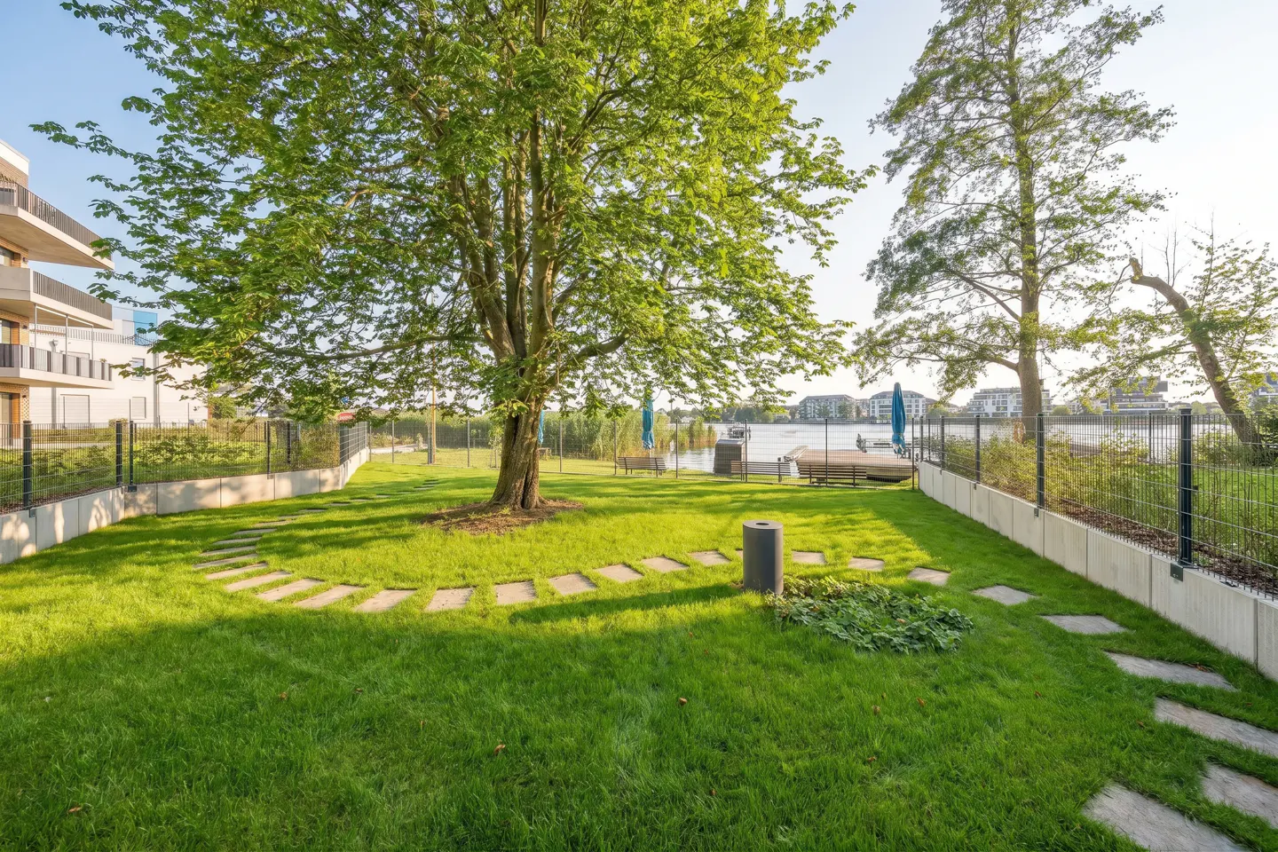 A lush green lawn with stone pavers leads to a waterfront area with benches and blue umbrellas. A large tree provides shade.