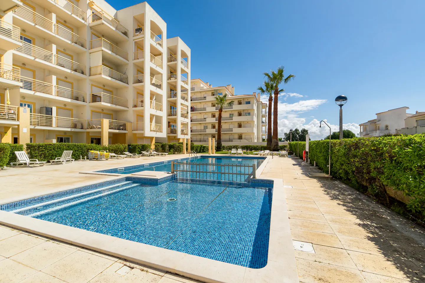 Exterior view of a blue tiled pool with apartment buildings and palm trees under a blue sky.