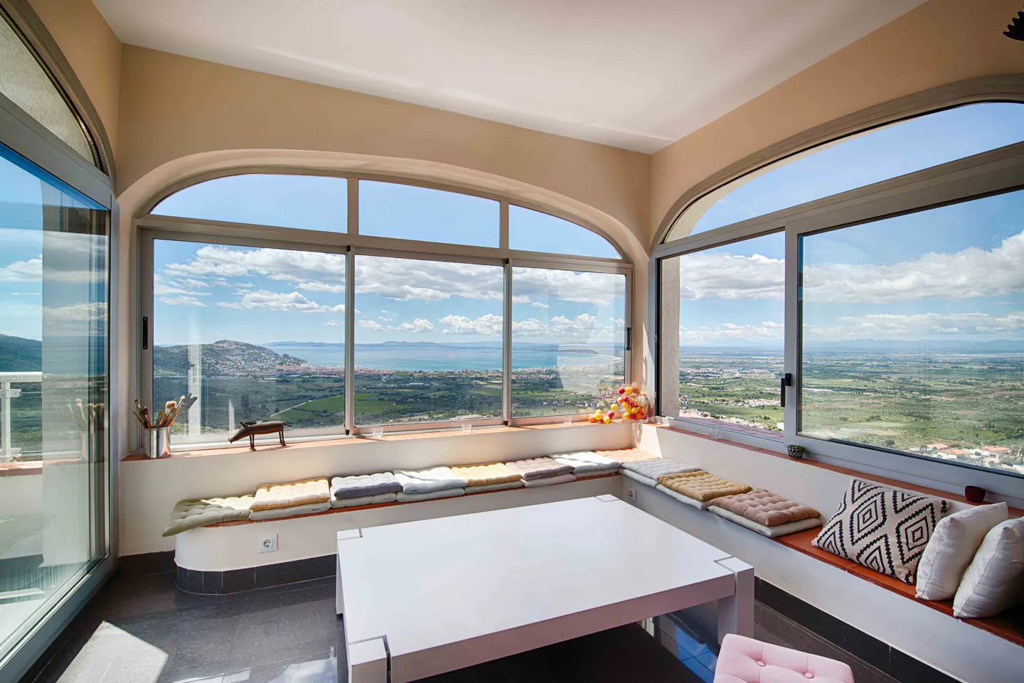 Bright sunroom with large arched windows overlooking a scenic landscape. Cushioned seating lines the windows, with a white table in the center.