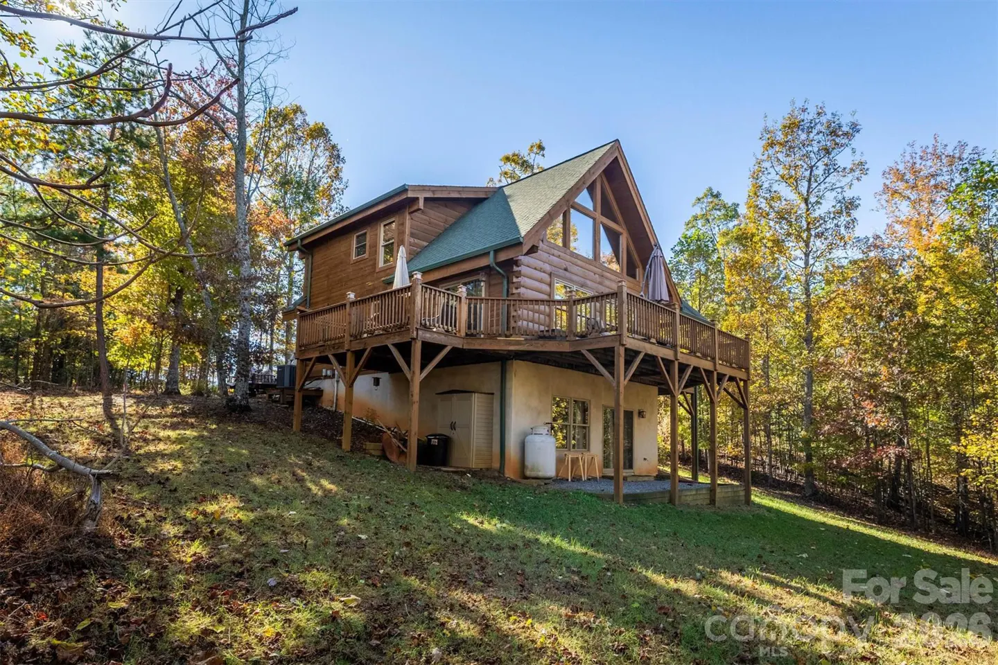 Exterior view of a two-story log cabin with a green roof and a large wooden deck, surrounded by trees with autumn foliage.