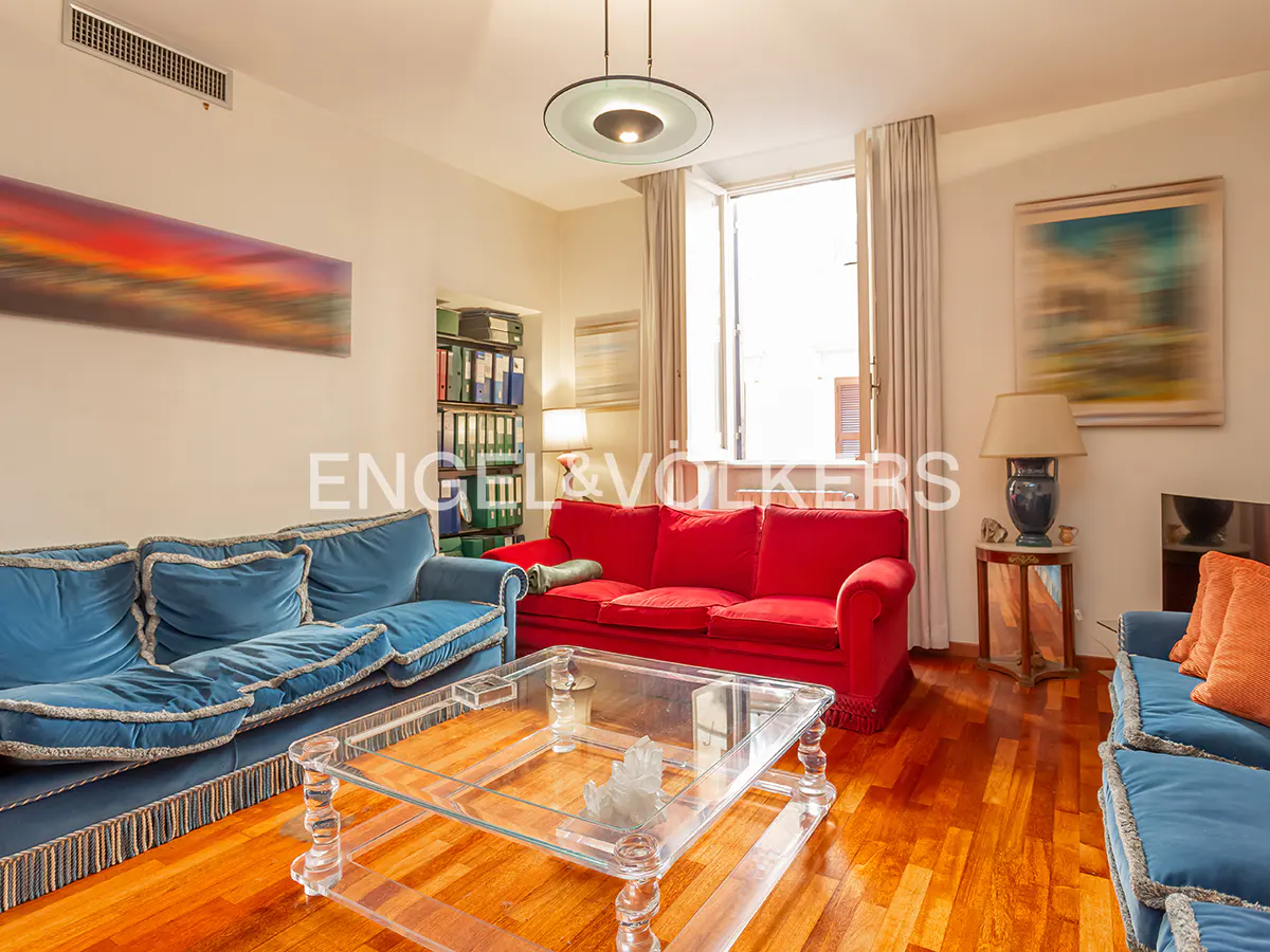 Living room with blue and red sofas, a glass coffee table, and hardwood floors. A window provides natural light.