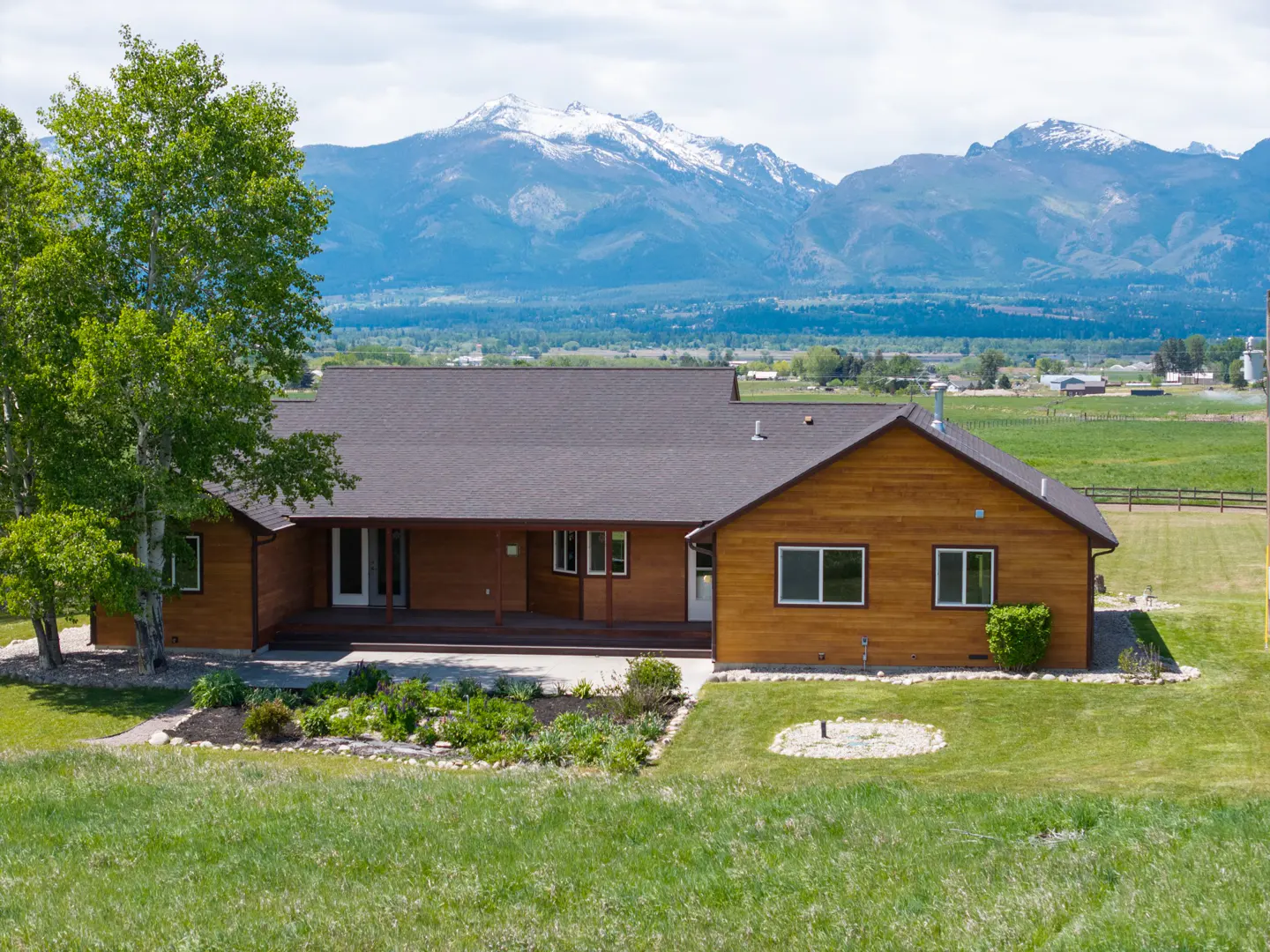 Single-story wood-sided home with a dark roof, front porch, and garden, set against a backdrop of snow-capped mountains.