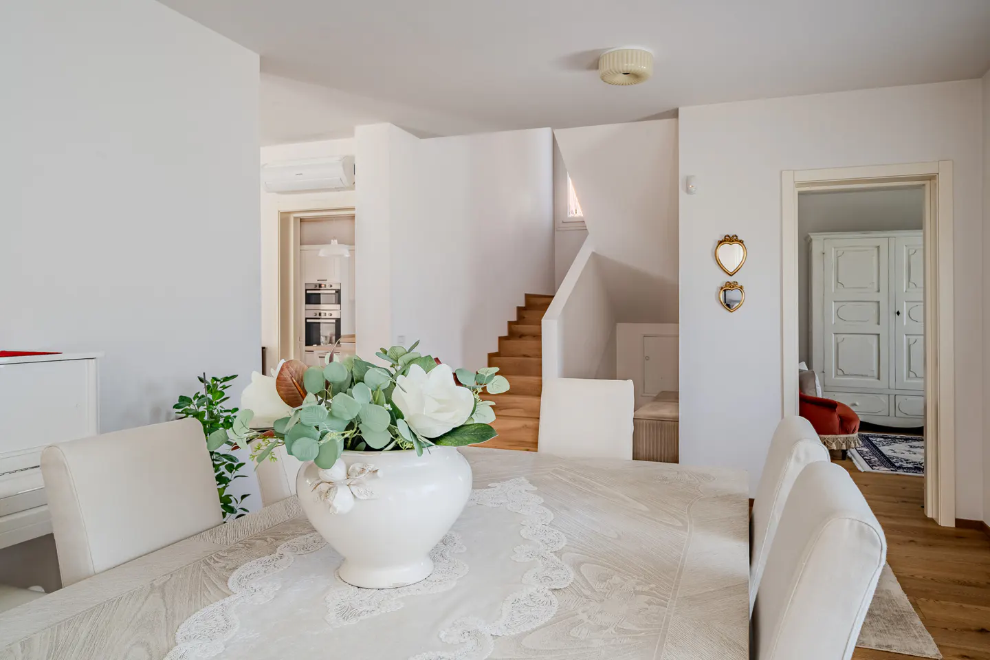 Bright, airy dining room with white walls, wood floors, and stairs. A white vase with flowers sits on a table with white chairs.
