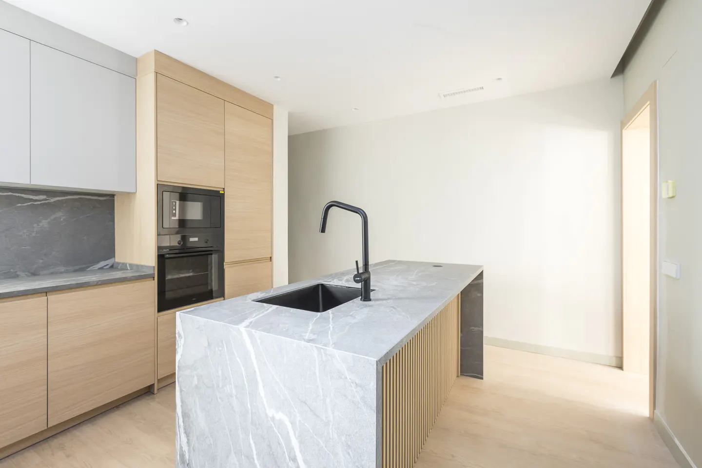 A modern kitchen with light wood cabinets, a marble island with a black sink and faucet, and light wood floors.