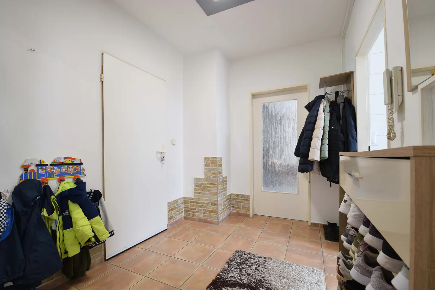 Entryway with white walls, terracotta tile floor, and a gray rug. Coats hang near a white door and a shoe rack.