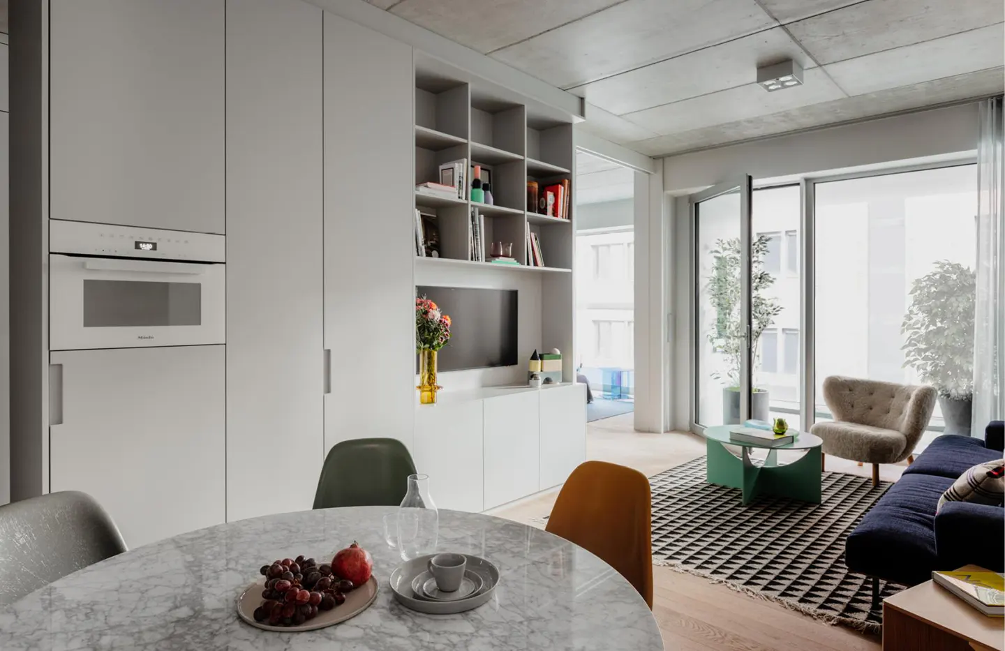 Modern apartment interior with a marble dining table, white cabinetry, open shelving, and a living area with a dark sofa.