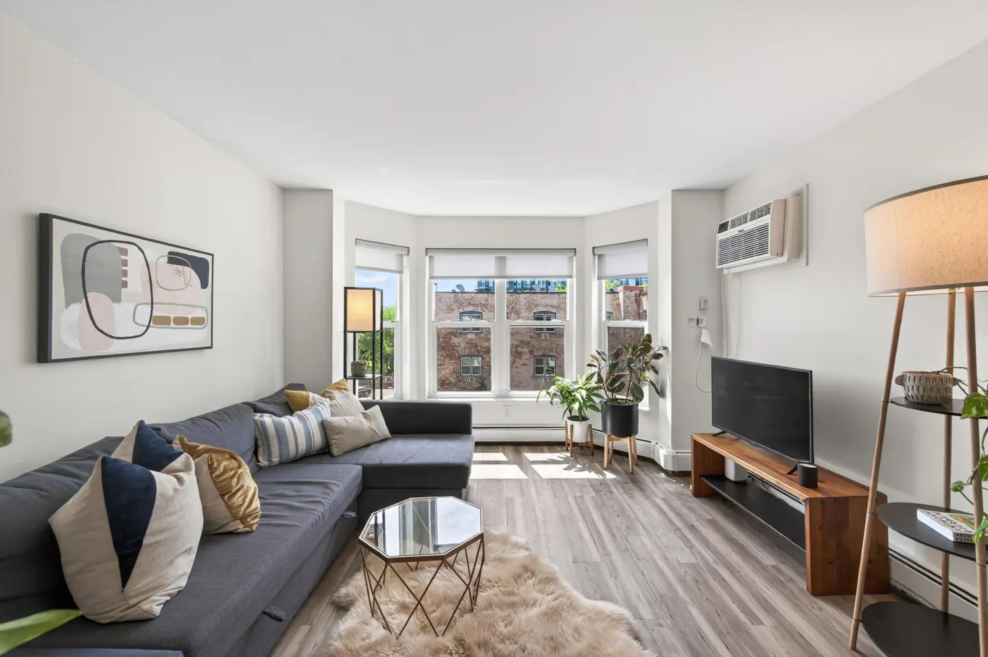 Bright living room with gray sectional sofa, geometric coffee table, and wood-look floors. Large windows offer natural light.