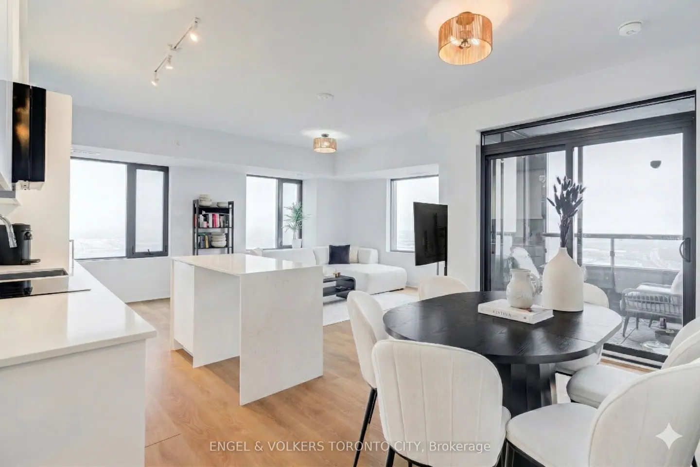 Bright, modern condo interior with white kitchen, island, and living area. Black dining table with white chairs. Balcony with sliding glass doors.