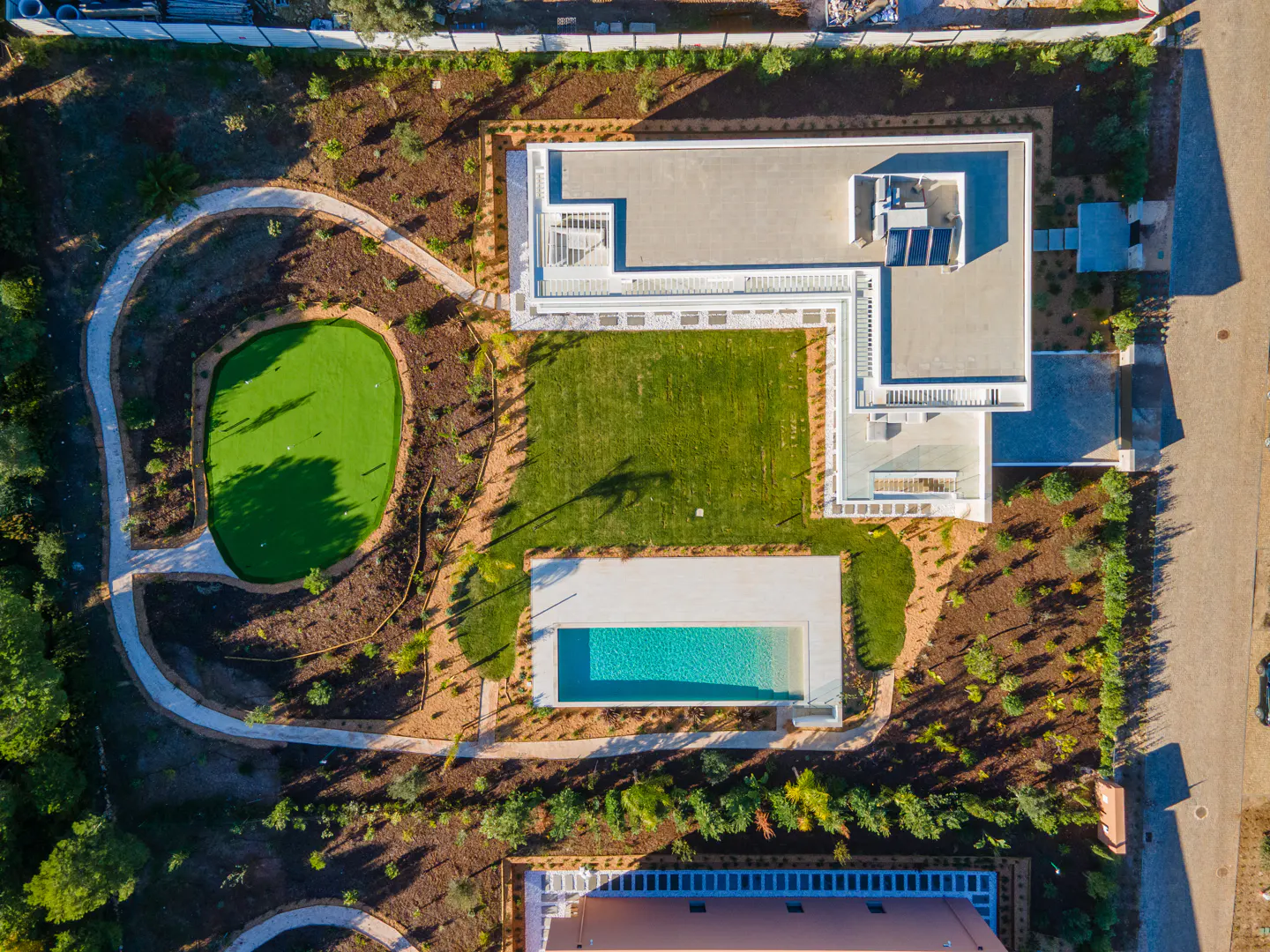 Aerial view of a modern white house with a pool, green lawn, and putting green, surrounded by trees and landscaping.