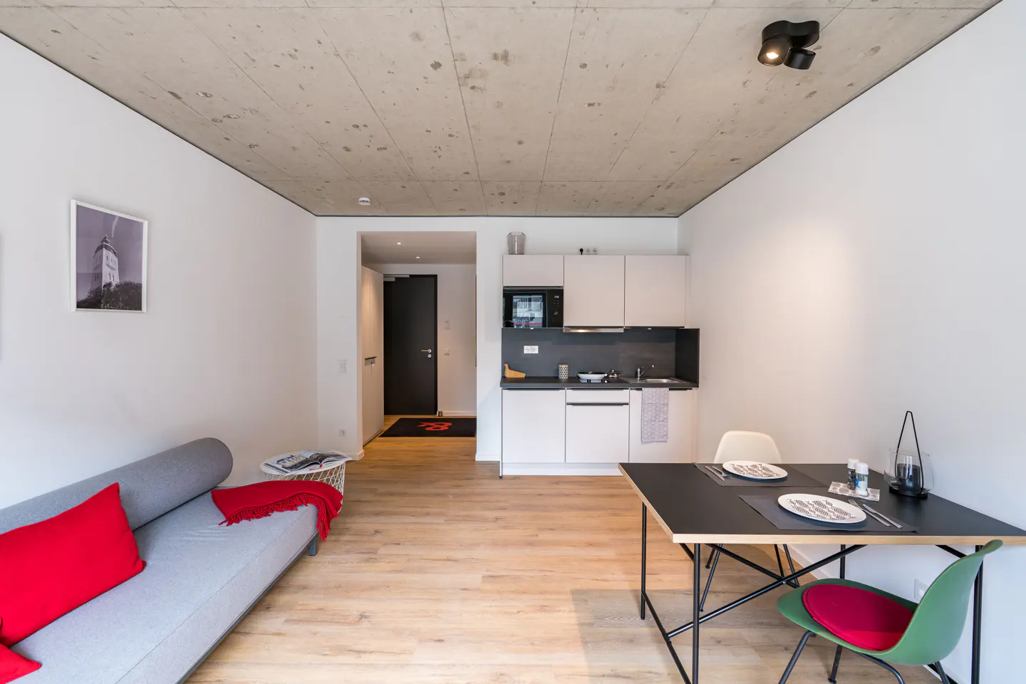 Studio apartment with a gray sofa, red pillows, a black table set for two, and a white kitchen against white walls.