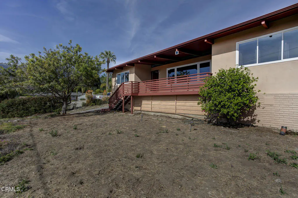Exterior view of a single-story home with a red deck and stairs, set on a dry, grassy hill under a blue sky.