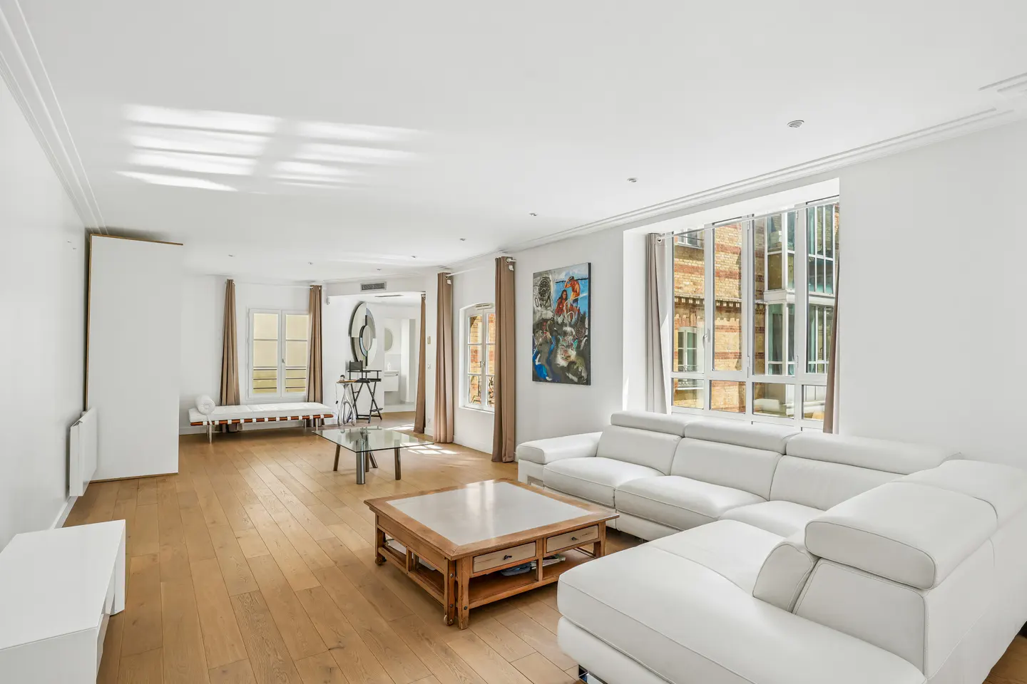 Bright, modern living room with hardwood floors, white walls, and a large white sectional sofa. Windows offer views of a brick building.