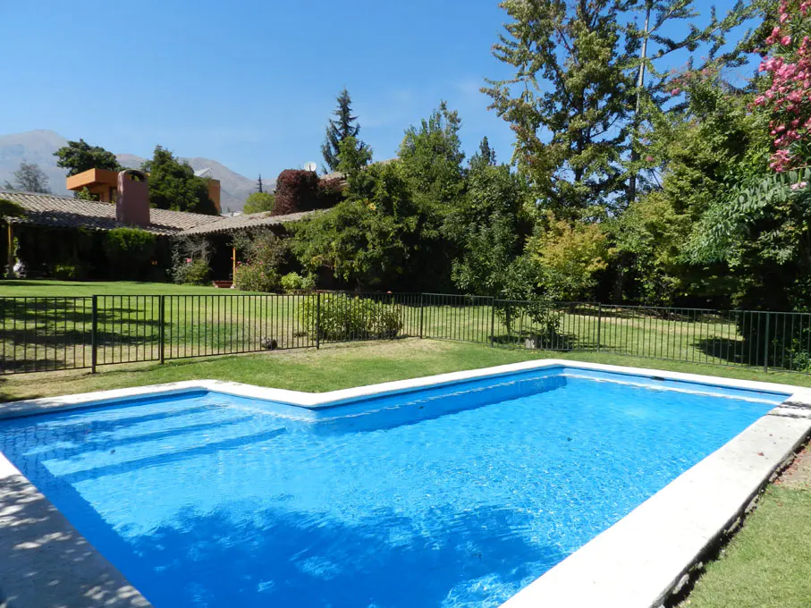A bright blue swimming pool is surrounded by green grass, trees, and a black metal fence. A house is visible in the background.