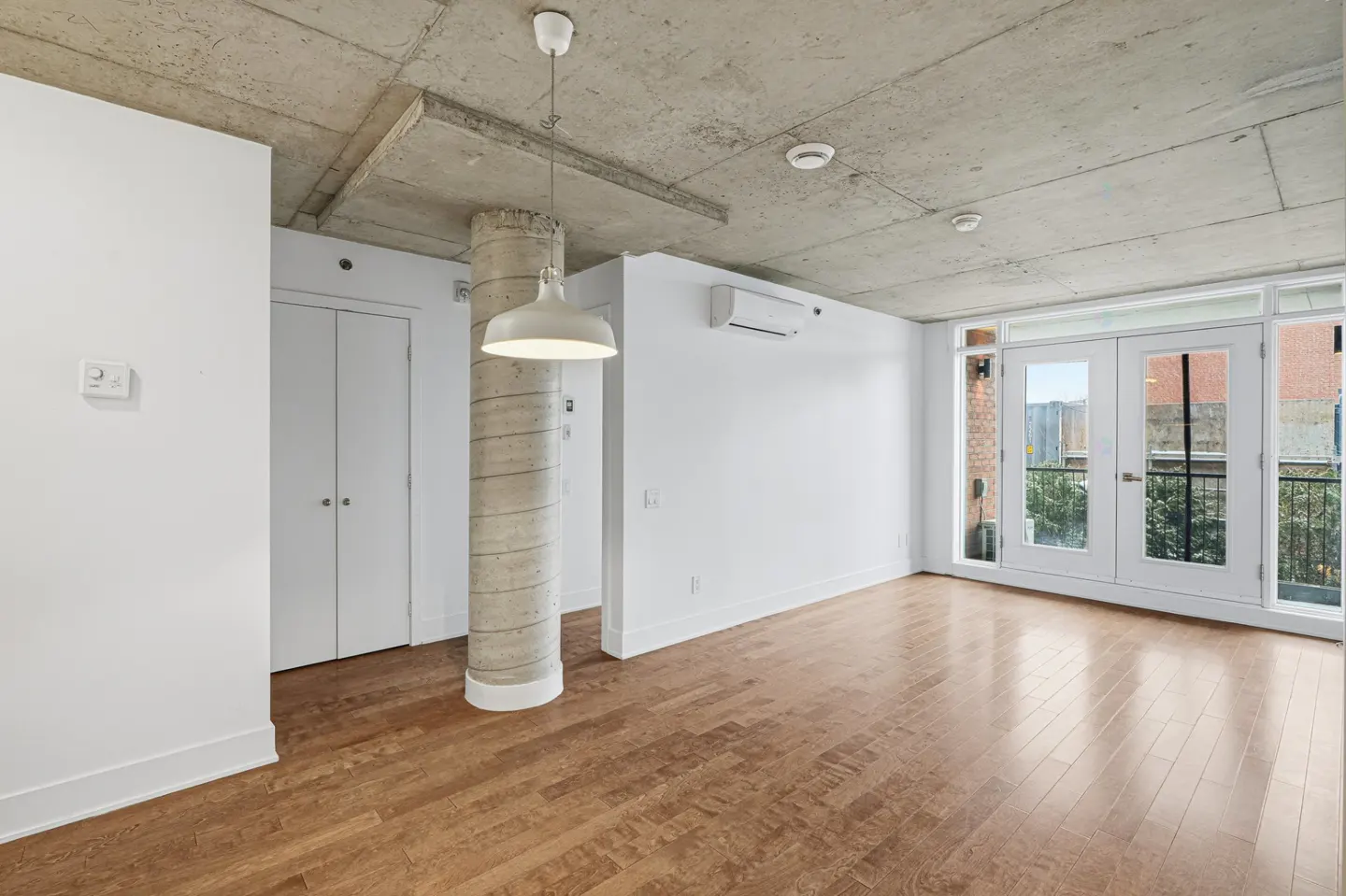 An empty room with hardwood floors, white walls, and a concrete ceiling. A white pendant light hangs near a concrete pillar. Double doors lead to a balcony.