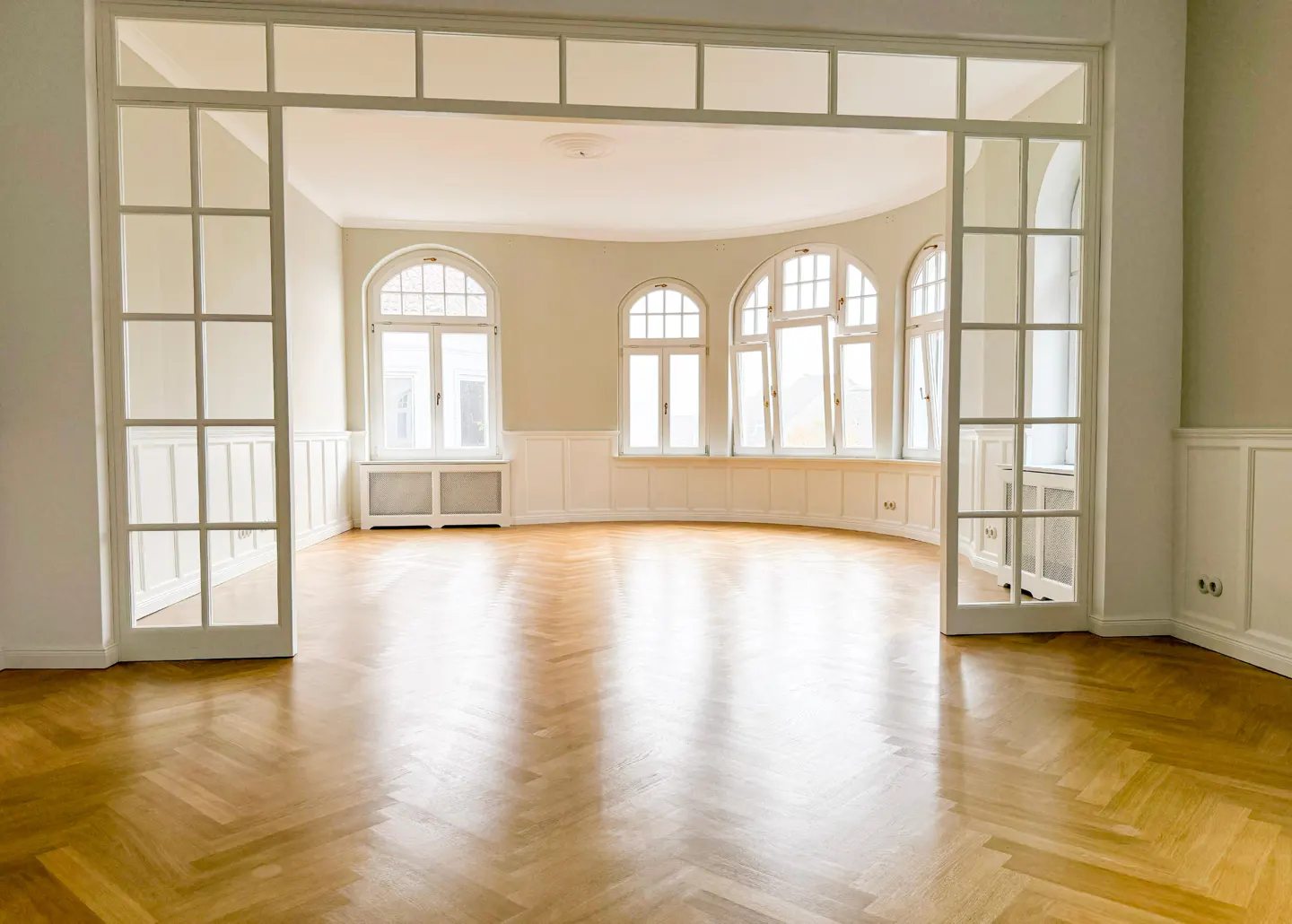 Bright, empty room with herringbone wood floors, white walls, and arched windows. A white, glass-paneled doorway leads into the space.