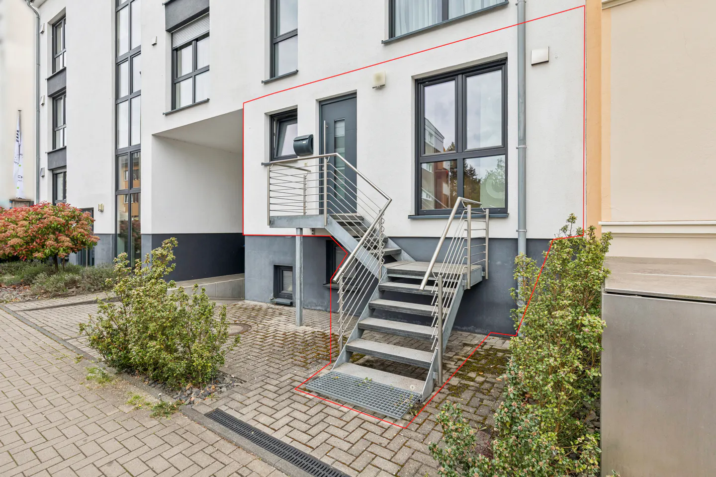 Exterior view of a white building with a metal staircase leading to a gray door, surrounded by greenery and a brick walkway.