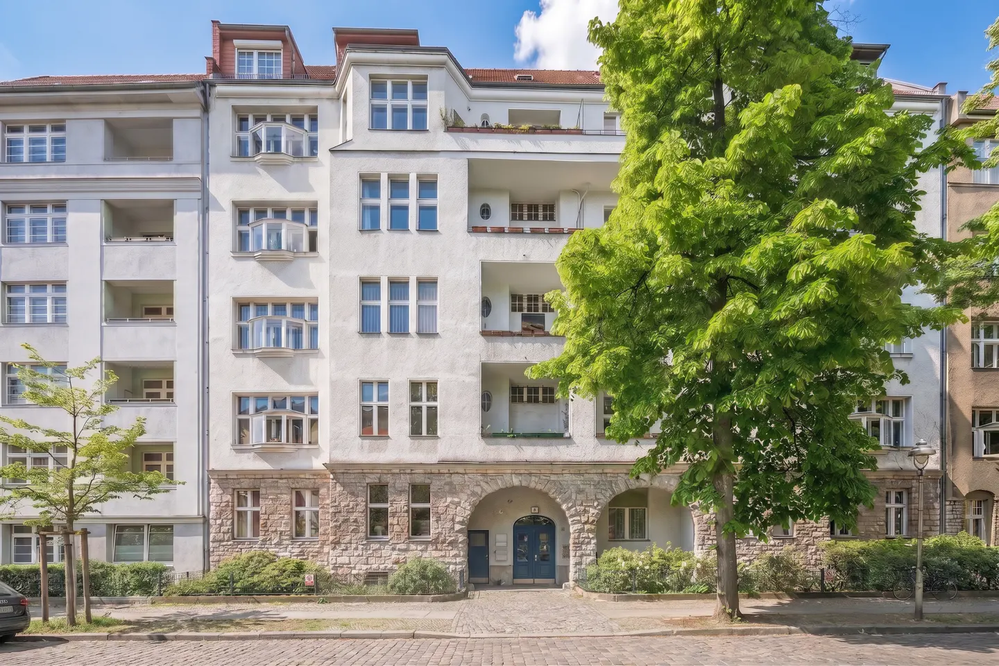 Exterior of a white apartment building with stone accents and blue doors, framed by green trees.
