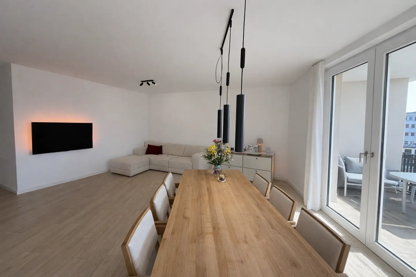 Bright, modern living room with a long wooden table, chairs, and a white sofa. Black pendant lights hang above the table. Balcony access visible.