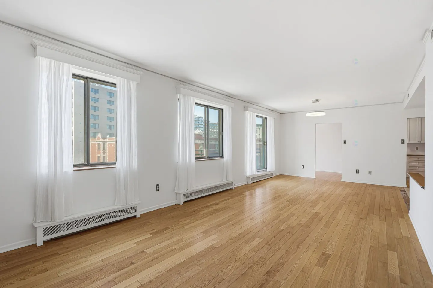 Bright, empty room with hardwood floors, white walls, and three windows with sheer white curtains. A glimpse of a kitchen is visible.
