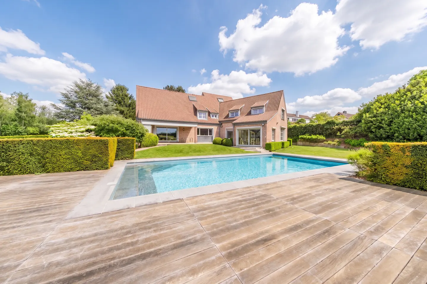 Backyard view of a brick house with a pool and wooden deck under a blue sky.