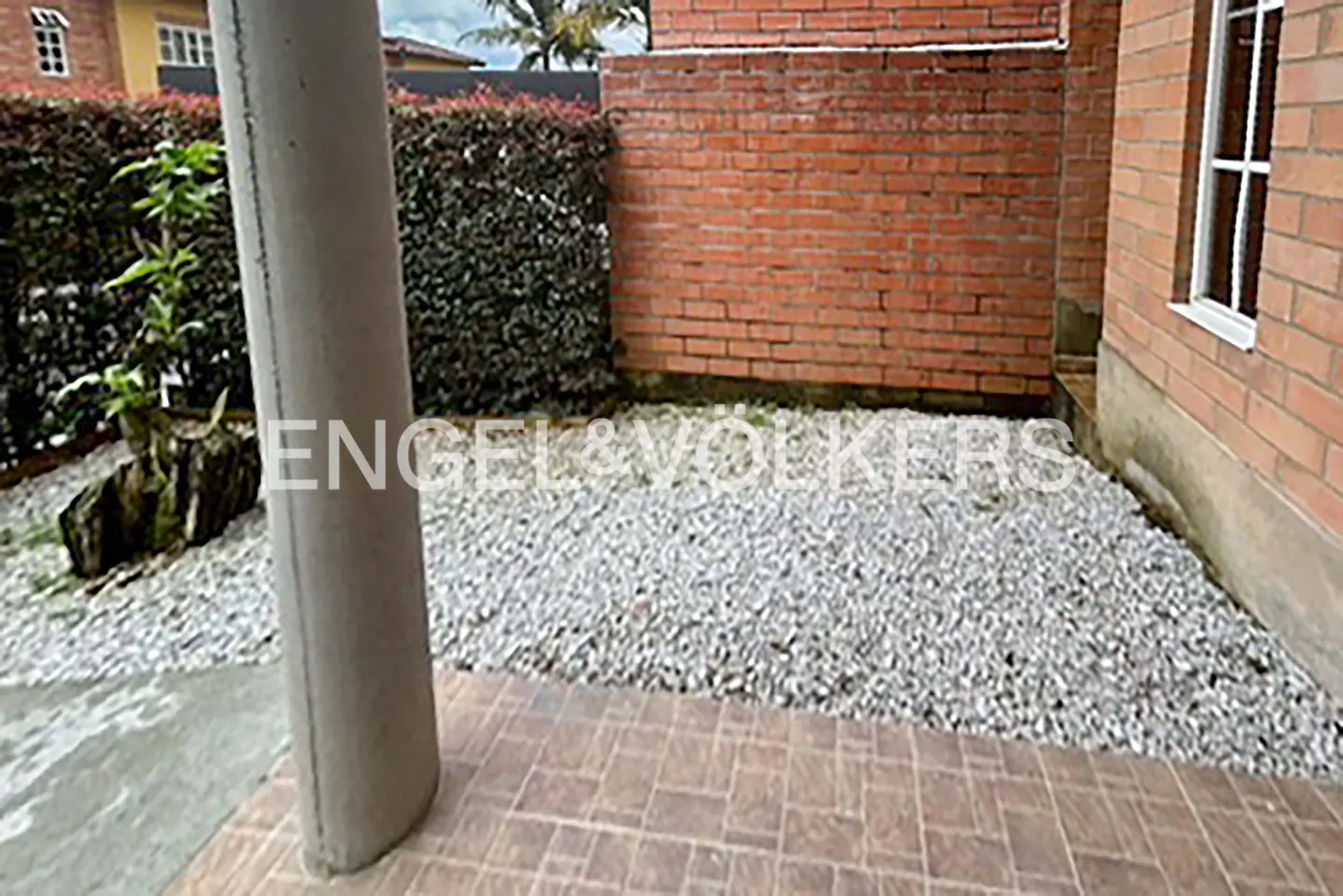 A patio area with gray gravel, brick walls, and a pillar. A green hedge is in the background. The Engel & Volkers logo is superimposed.