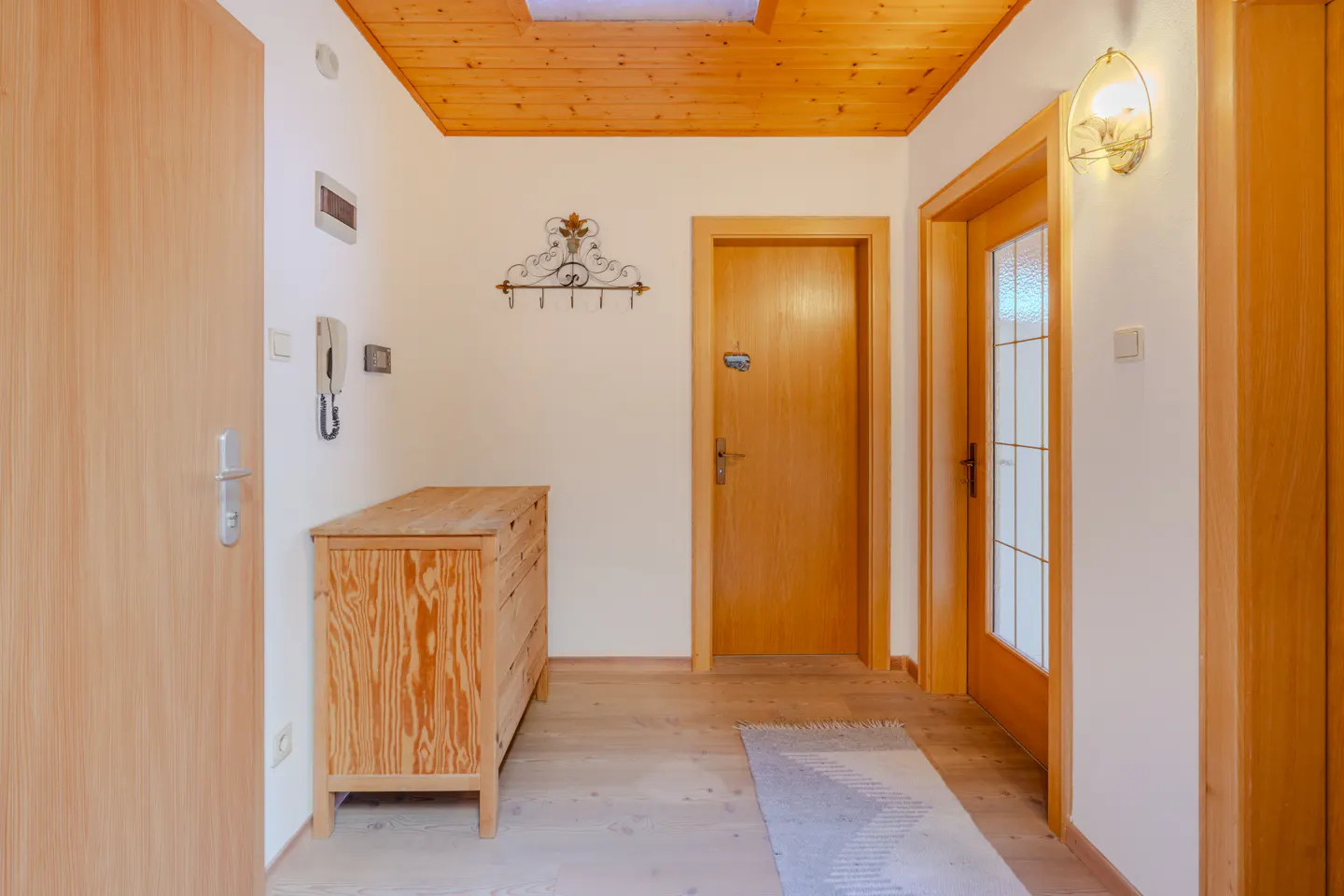 Entryway with wood floors, doors, and ceiling. A wooden dresser sits near a wall with a coat rack and intercom. A rug leads to a glass-paneled door.