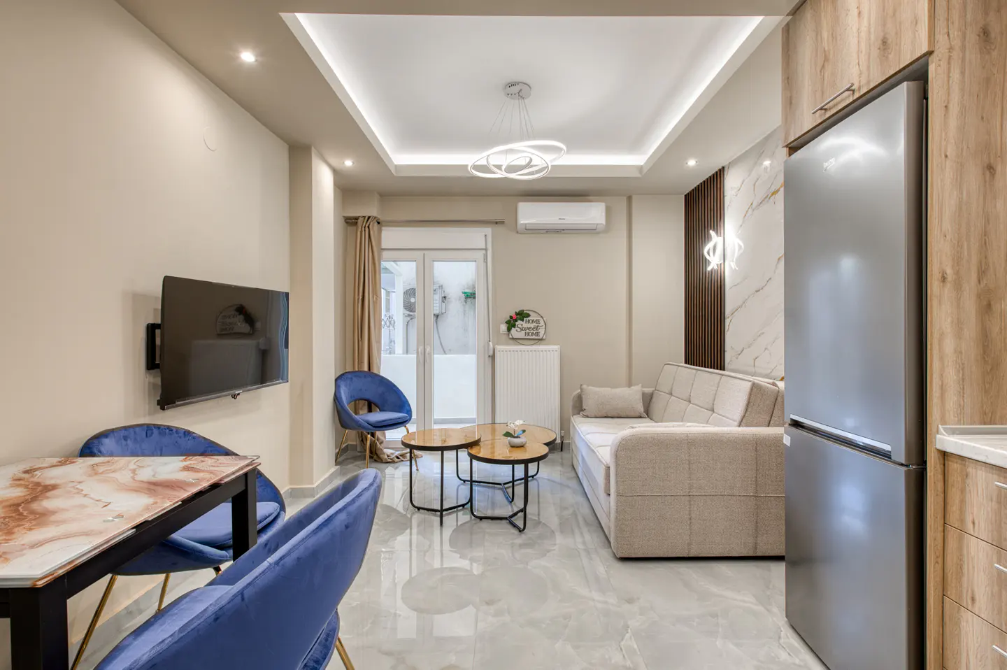 A modern apartment living room with blue chairs, a marble table, a beige sofa, and a stainless steel refrigerator.
