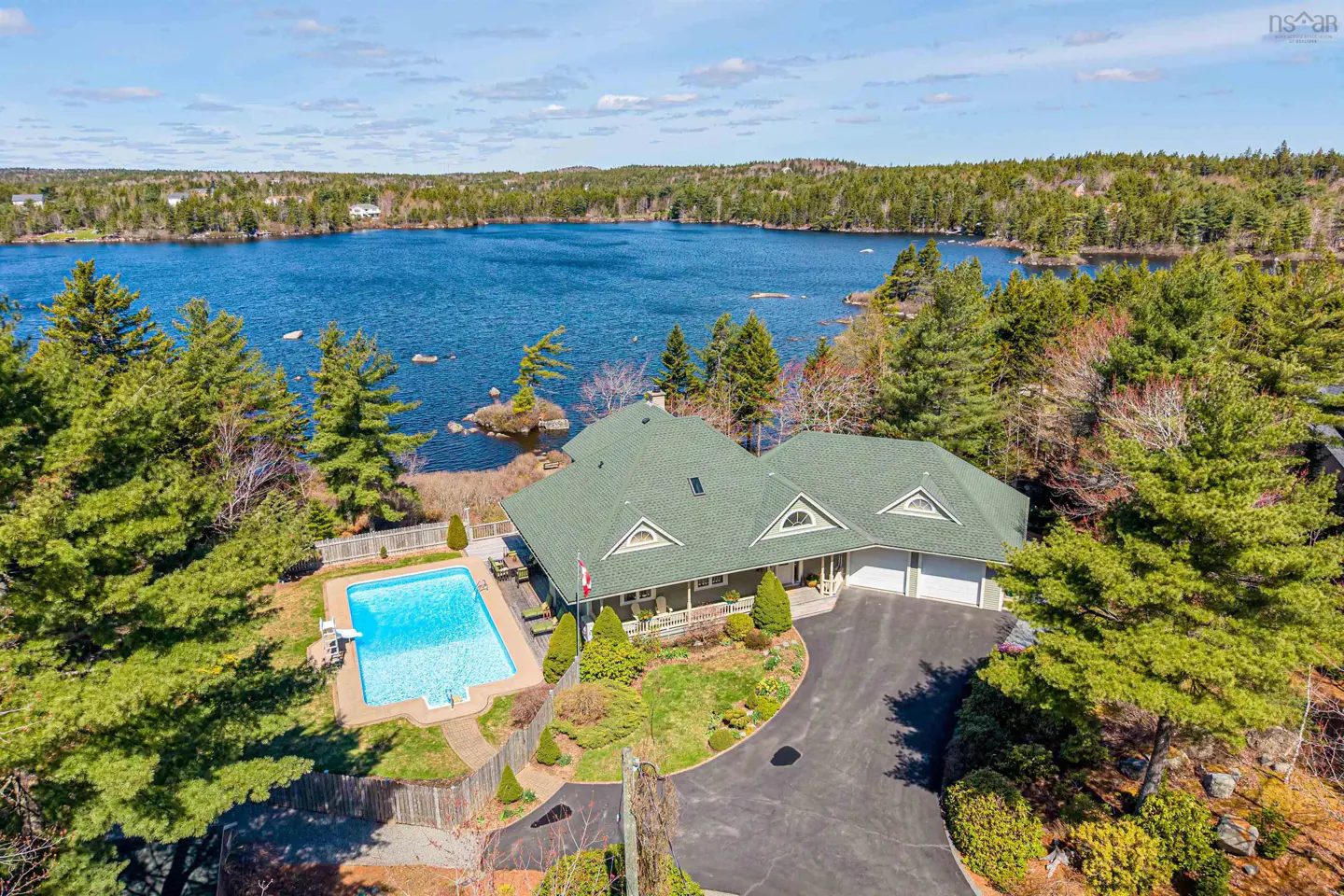 Aerial view of a lakefront home with a green roof, pool, and a paved driveway surrounded by trees.