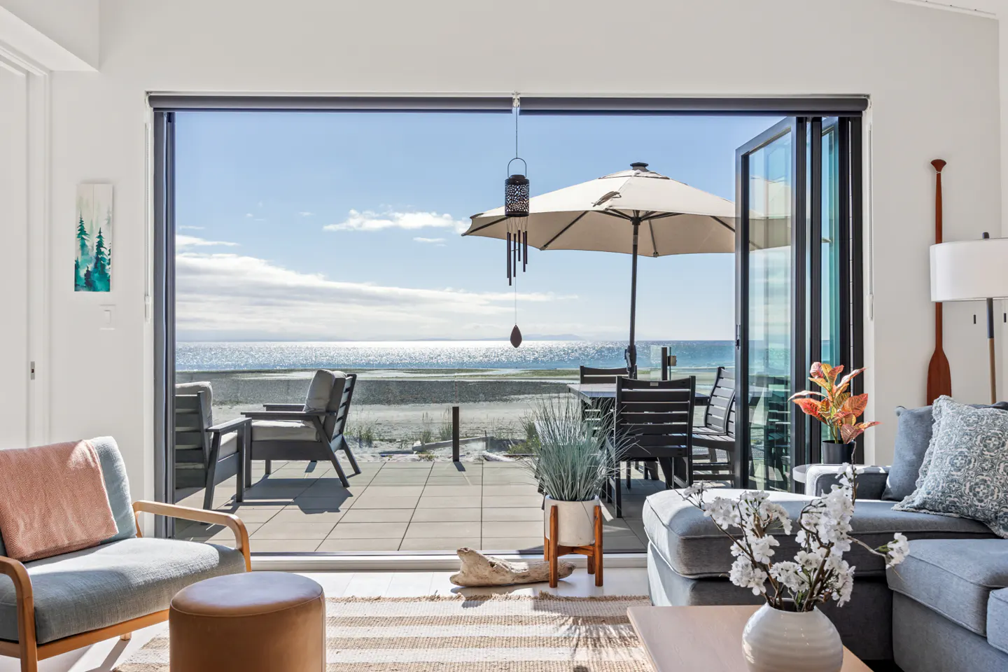 Living room with open glass doors to a patio overlooking a beach and ocean on a sunny day.
