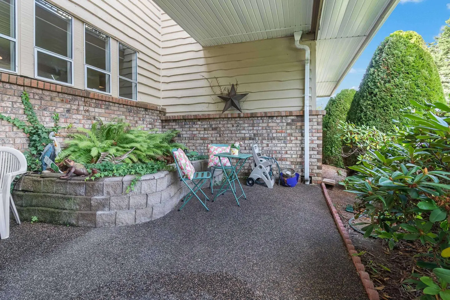 Covered patio with brick wall, garden bed, and green metal table and chairs. A star decoration hangs on the beige siding.