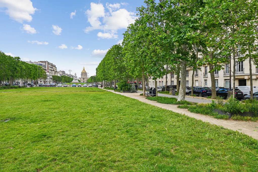 Paris park view with green grass, trees, and buildings. The Invalides dome is visible in the distance. Cars are parked along the street.
