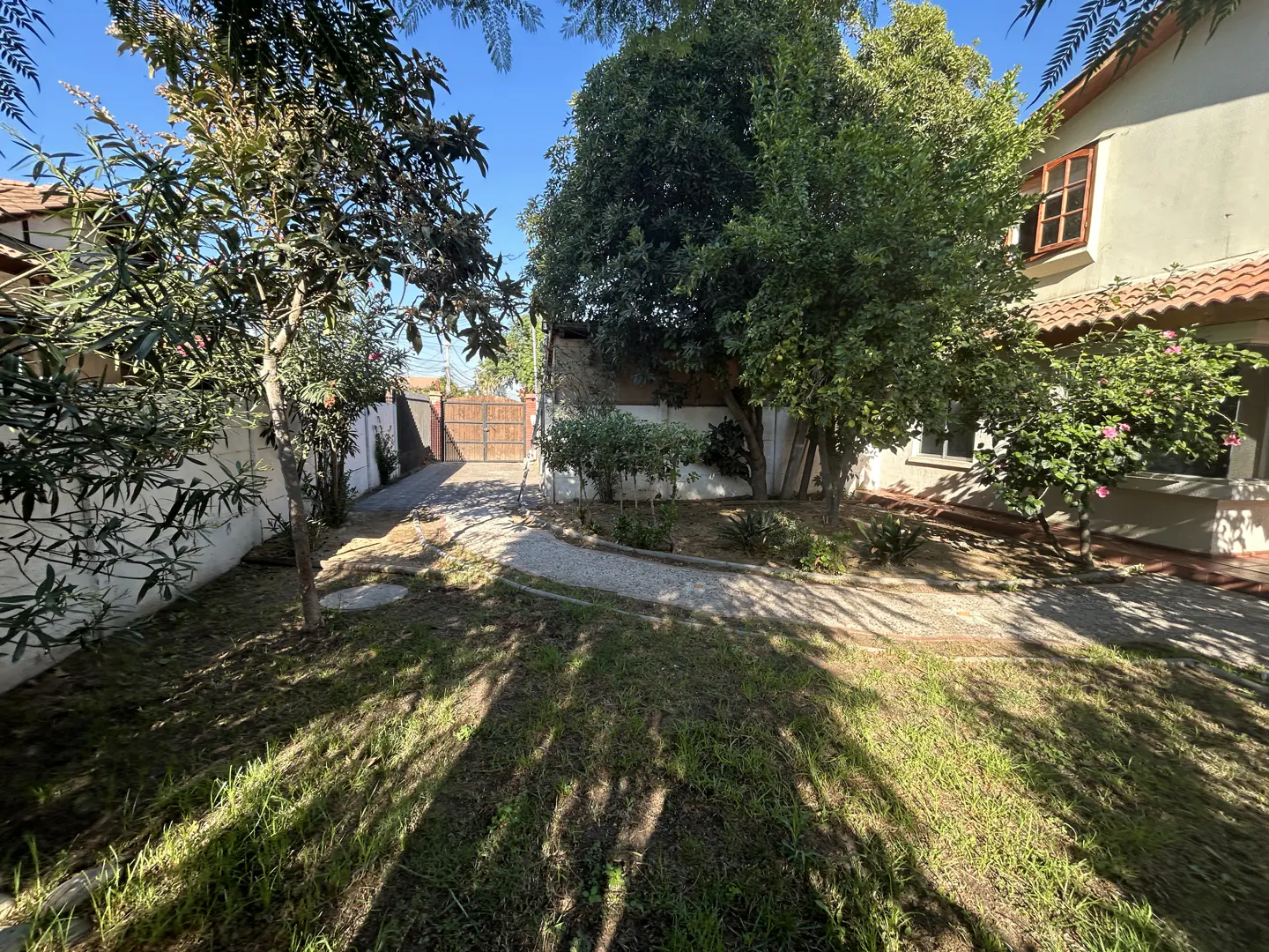 A sunny front yard with green grass, trees, and a gravel driveway leading to a wooden gate. A house with a red tile roof is visible.