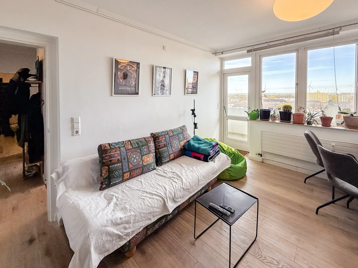 Bright living room with a couch, pillows, and a small table. Large windows offer a city view. Plants line the windowsill.