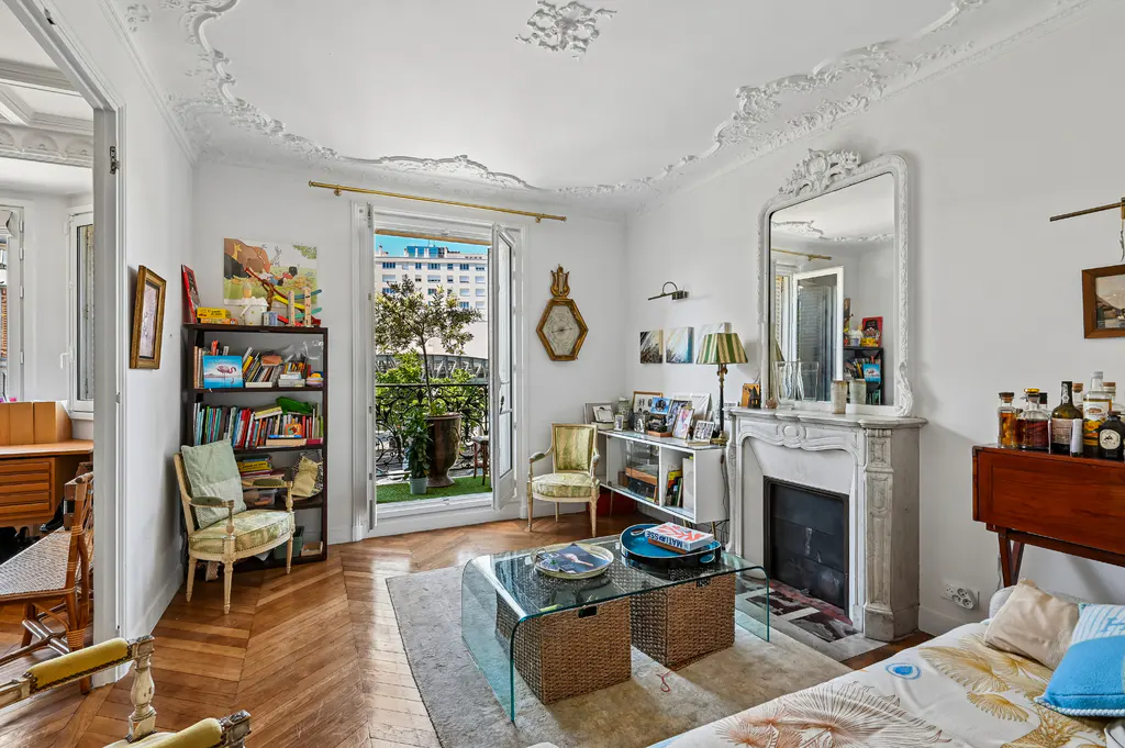 Bright living room with white walls, ornate ceiling, fireplace, and balcony with city view. Hardwood floors and glass coffee table.