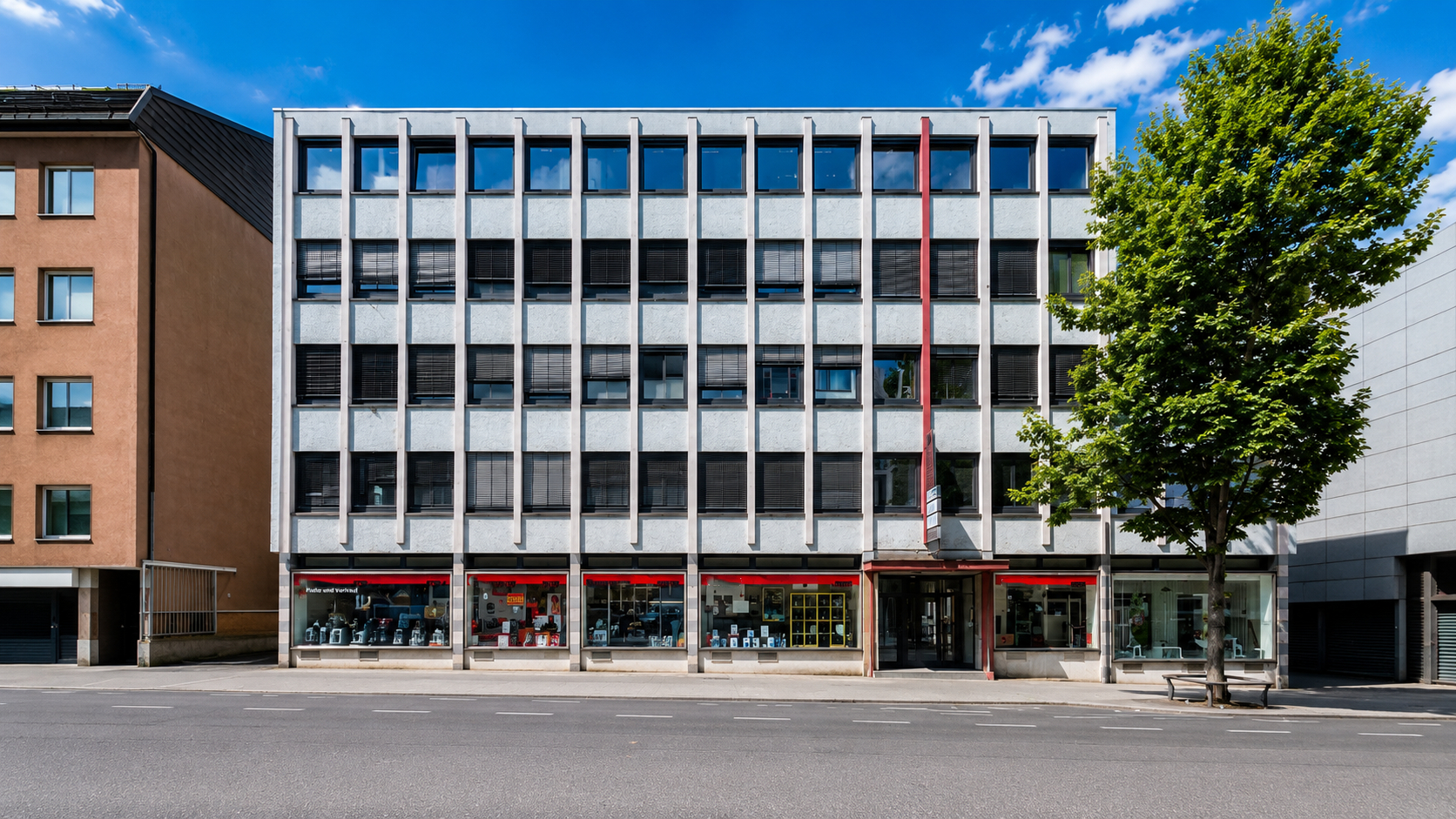 Modern four-story office building with a grid facade, ground-floor retail, and a large tree under a blue sky.