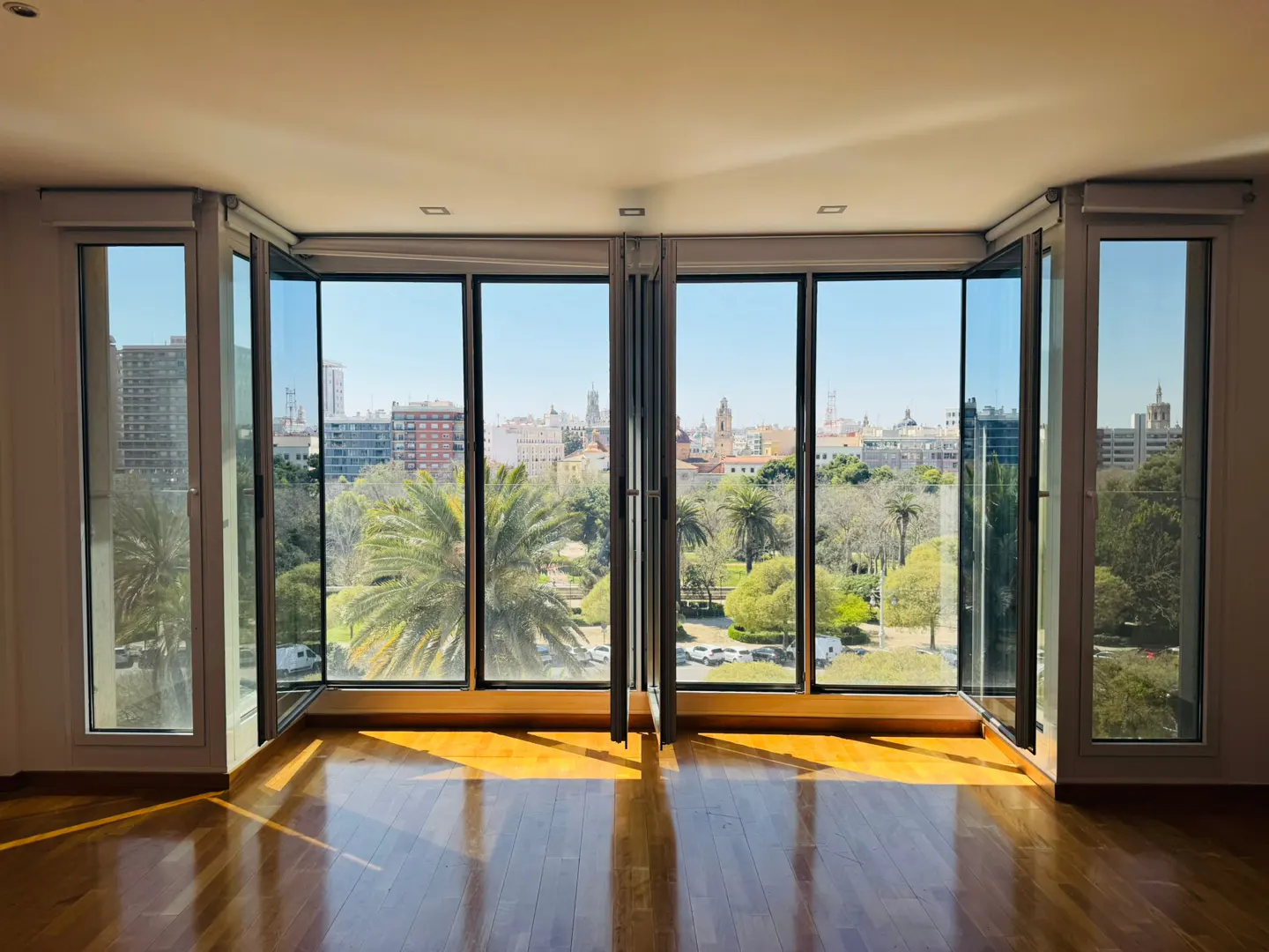 Bright room with open folding glass doors. Outside, a park with palm trees and city buildings under a blue sky. Sunlight reflects on the wooden floor.