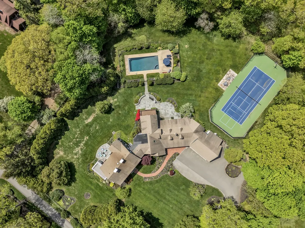 Aerial view of a large tan house with a pool, tennis court, and green lawn surrounded by trees.