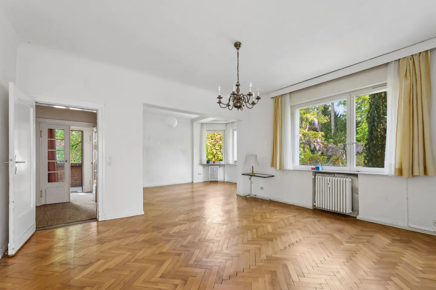 Bright, empty living room with herringbone wood floors, white walls, a chandelier, and large windows with yellow curtains.