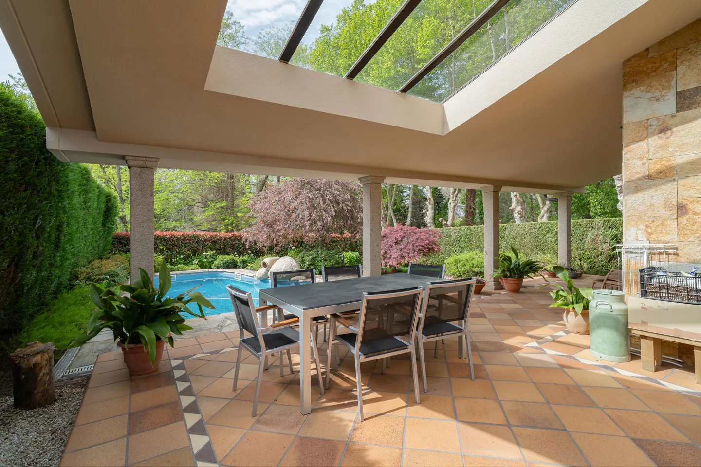 Covered patio with a table and chairs overlooks a pool and green garden. Stone pillars and a skylight add architectural detail.