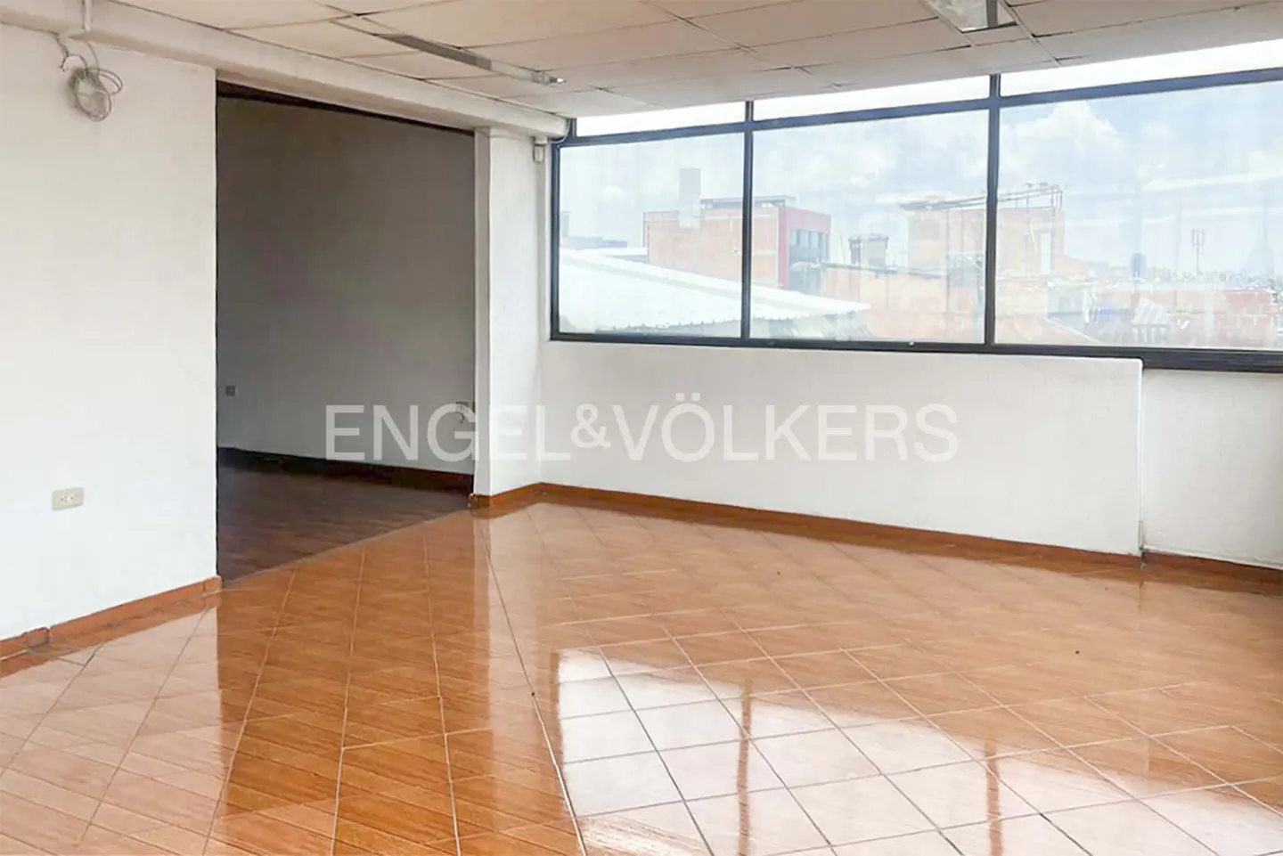 Empty room with brown tile floor, white walls, and large windows showing a city view. Engel & Völkers logo on the wall.