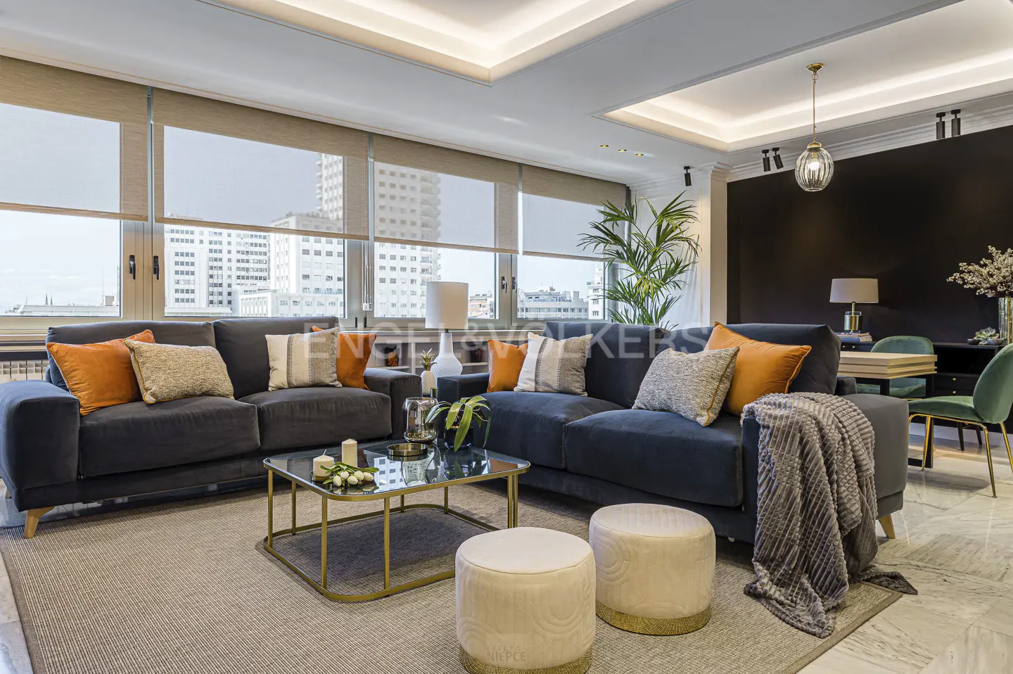 Living room with blue sofas, orange pillows, glass coffee table, and beige ottomans on a gray rug. Large windows with city views.