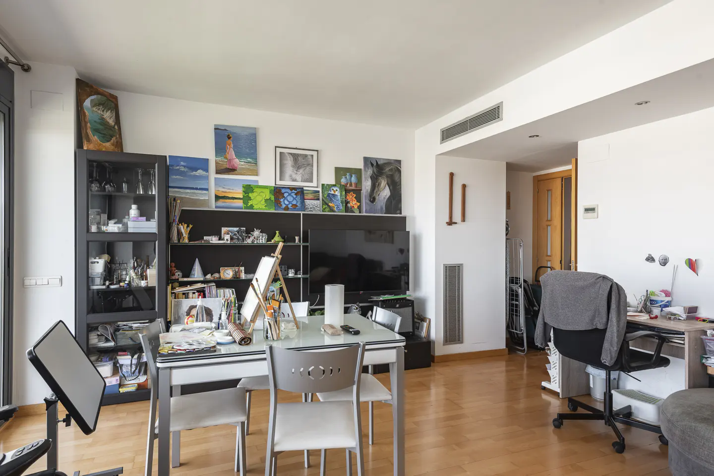 Bright living room with wood floors, white walls, and art. A glass table with chairs sits near a black bookcase and TV. Paintings adorn the wall above.