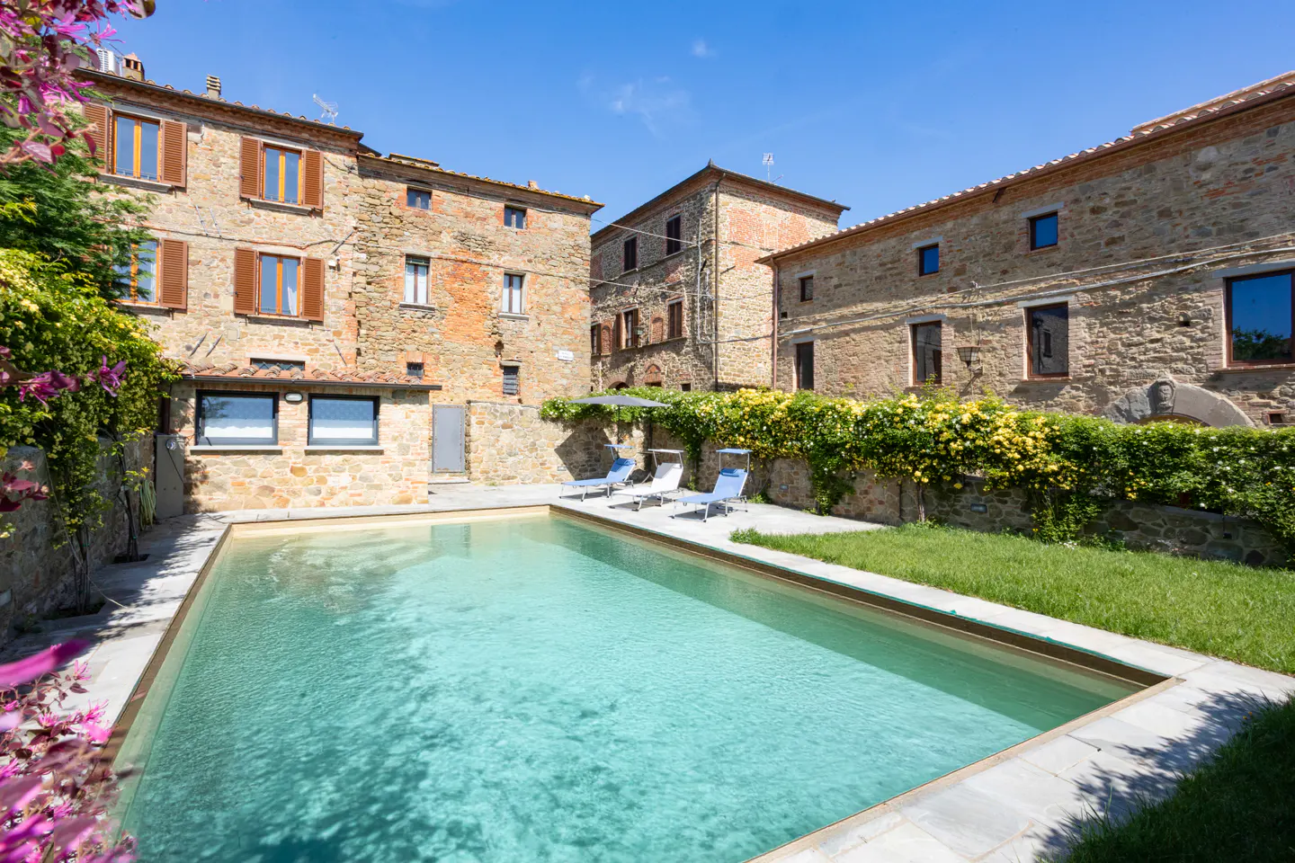Outdoor pool with lounge chairs surrounded by stone buildings and greenery under a blue sky.