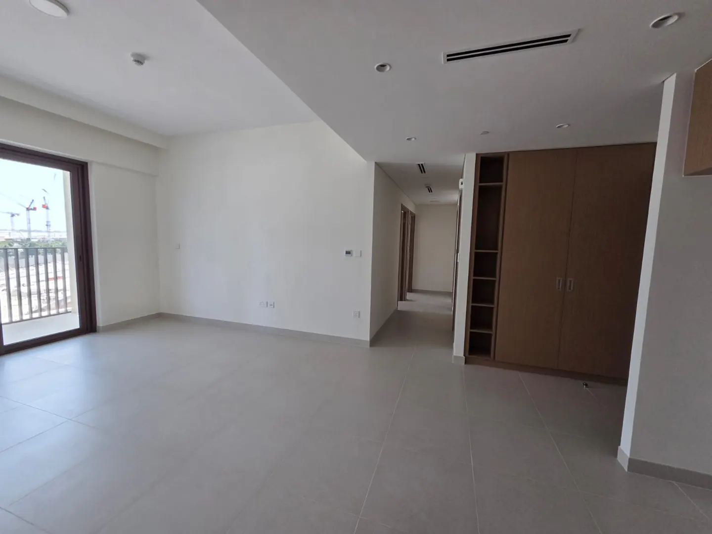 An empty room with gray tile floors, white walls, and a balcony with a view of construction cranes. A hallway leads to other rooms. A built-in closet is on the right.