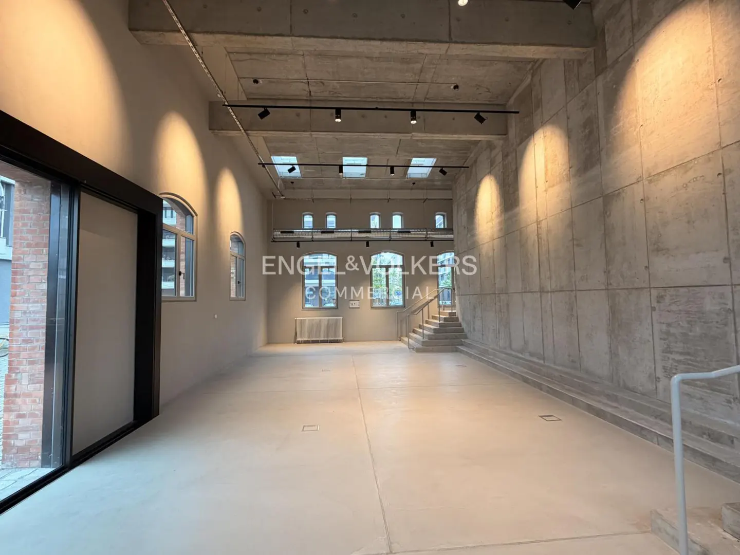 Bright, empty commercial space with concrete walls, track lighting, and arched windows. Stairs lead to an upper level. Engel & Völkers logo visible.