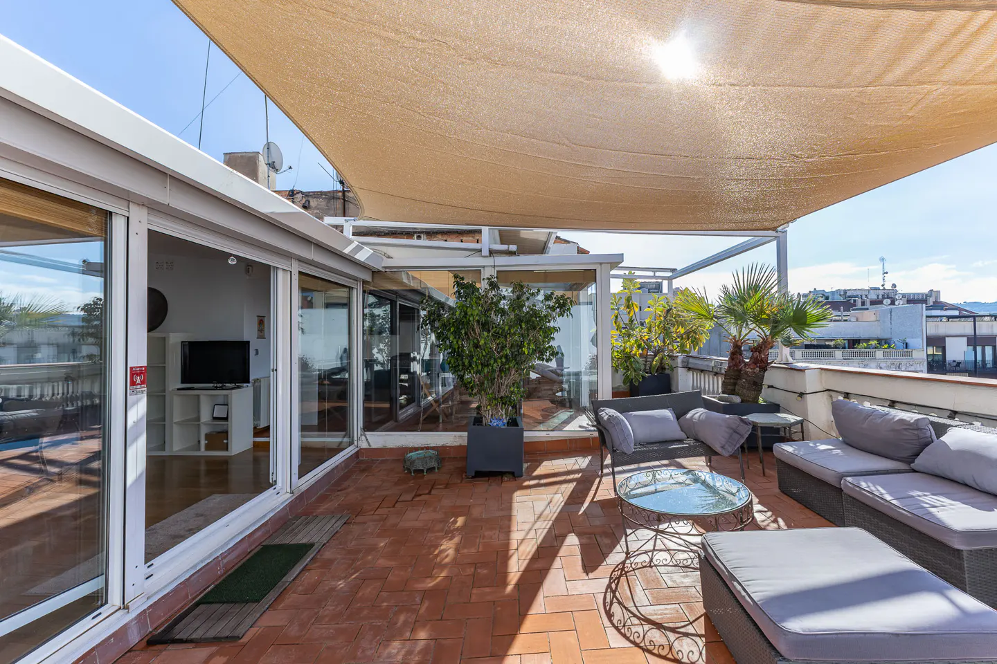 Rooftop terrace with brick flooring, gray wicker furniture, and potted plants under a beige sunshade. Sliding glass doors lead to an indoor living space.