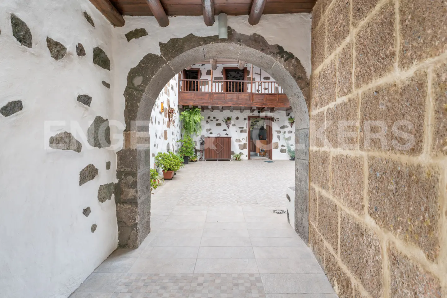 Stone archway leads to a courtyard with white walls, dark stones, and a wooden balcony.
