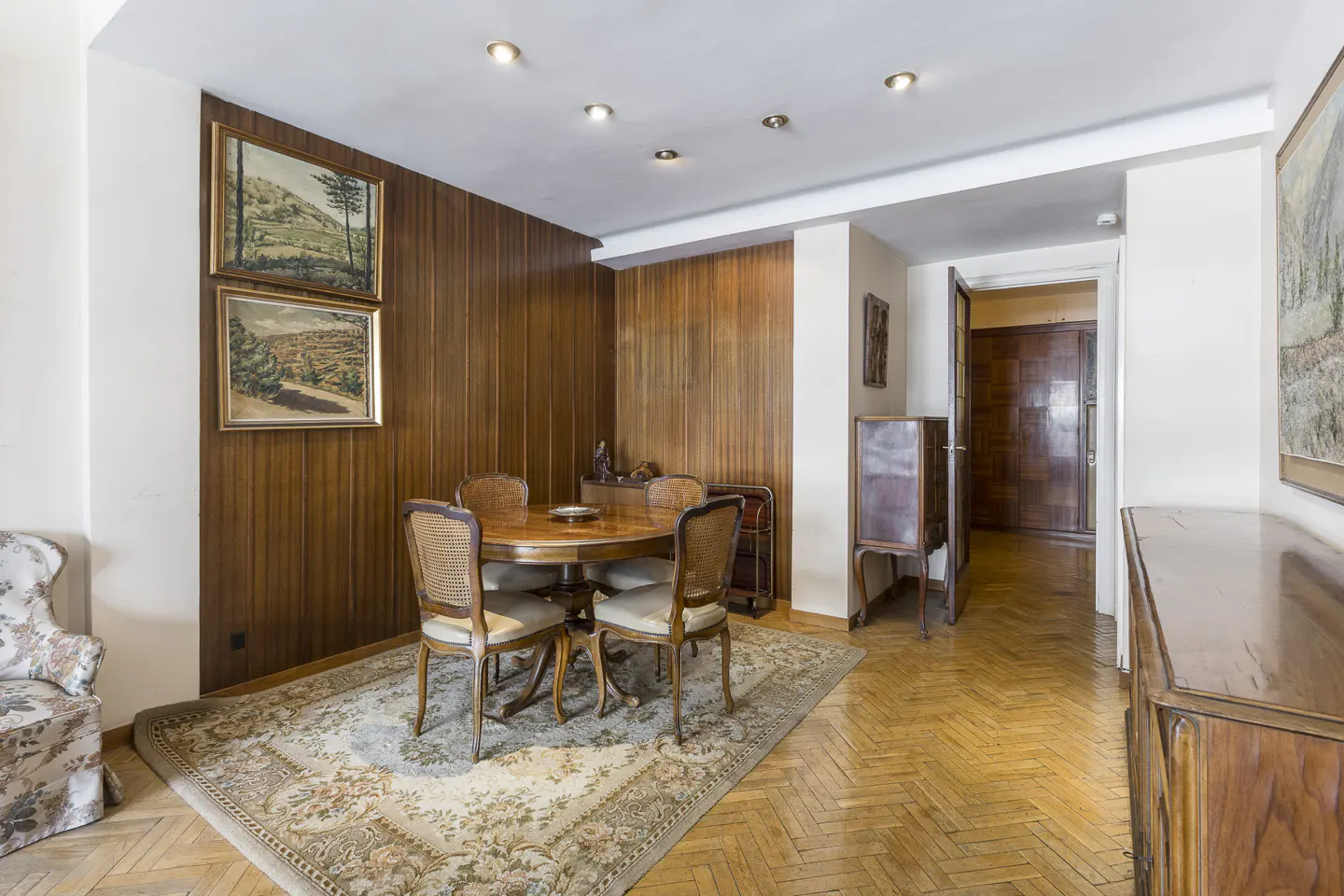 A dining room with wood-paneled walls, a round table with four chairs on a floral rug, and parquet floors.