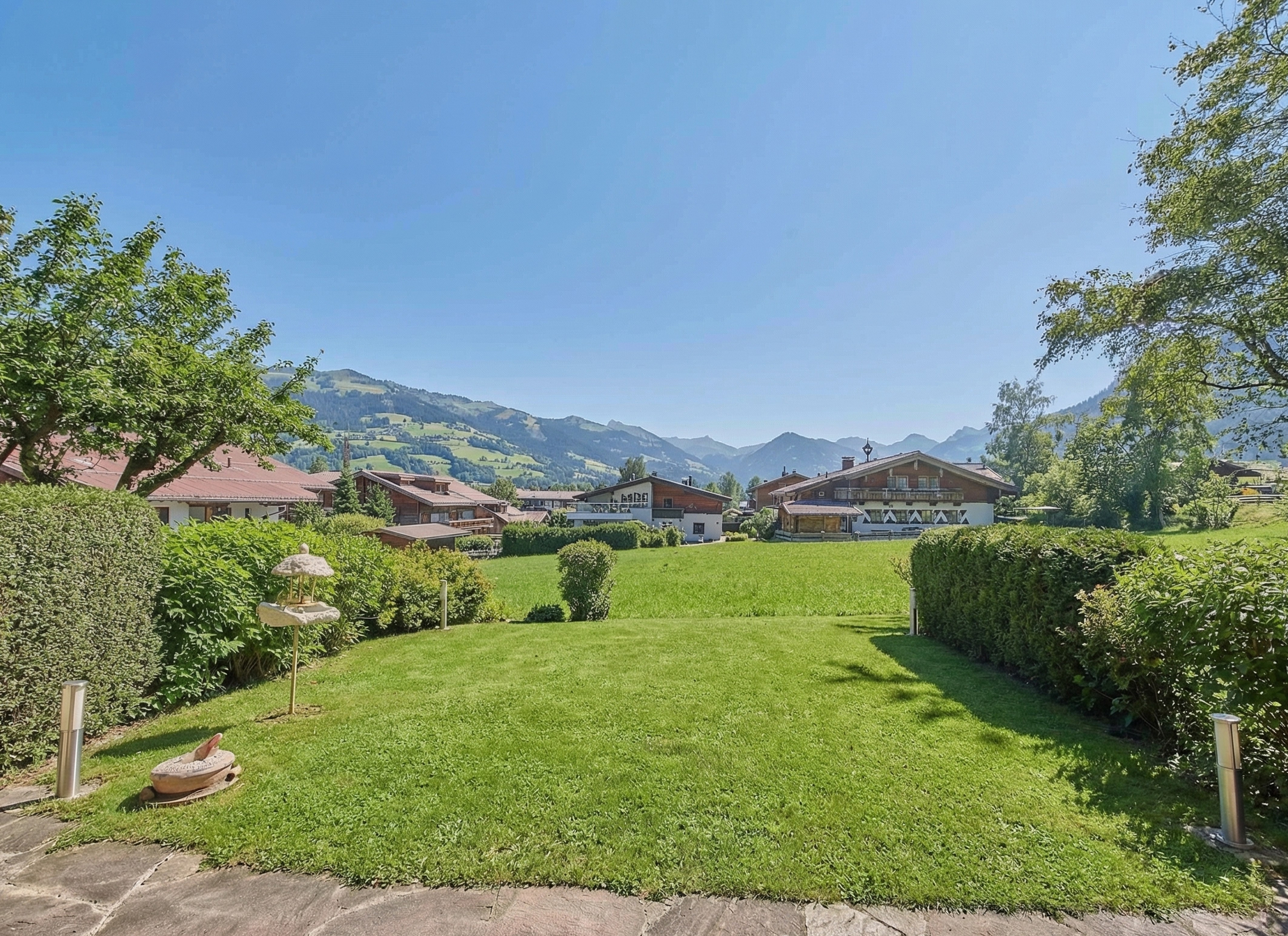 A lush green lawn framed by hedges leads to a scenic view of traditional alpine houses against a backdrop of mountains. [AI-generated text] [AI-modified]