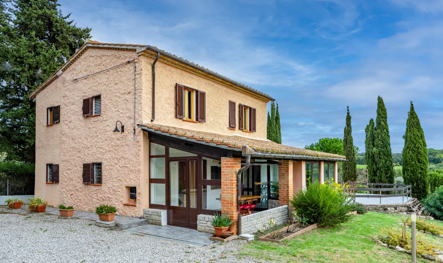 Two-story peach stucco house with brown shutters and a glass-enclosed porch. Tall cypress trees and a blue sky are in the background.